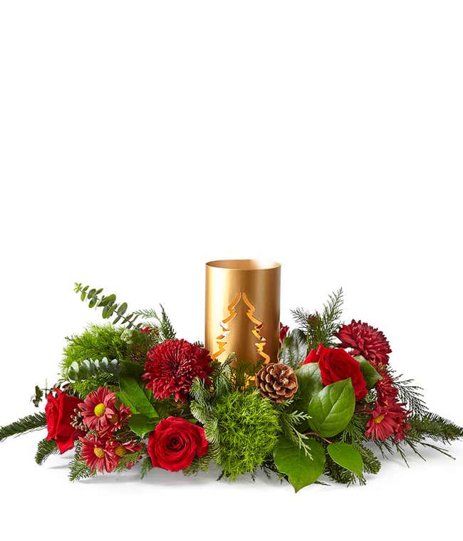 Holiday centerpiece featuring red roses, red poms, and red daisy poms surrounded by lush seasonal greenery and pinecones. Accented with a gold candle holder featuring a Christmas tree cutout