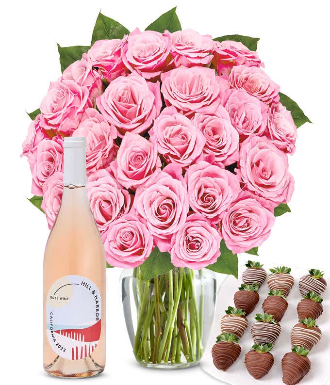 A bouquet of 2 dozen soft pink roses arranged in a clear glass vase is displayed beside a bottle of Hill & Harbor Rosé and a neat assortment of 12 chocolate-covered strawberries.