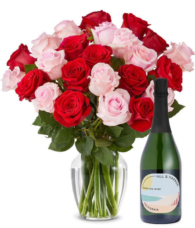 A mix bouquet of two dozen red and blush roses fills a clear glass vase and is paired with a bottle of Hill & Harbor California Sparkling Wine.