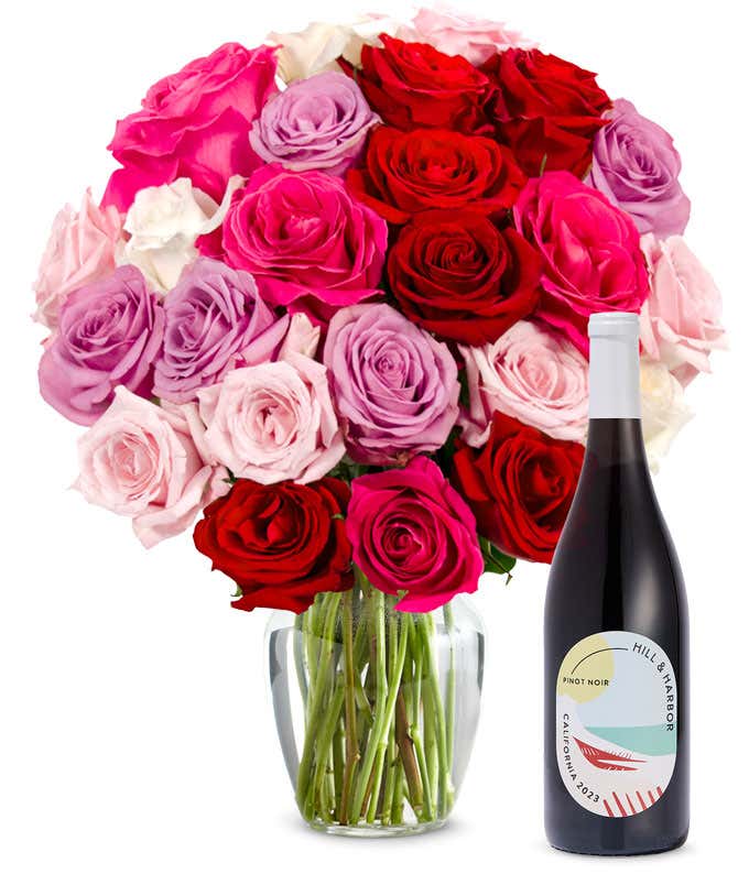 Two dozen pink, purple and red roses arranged in a clear glass vase sits beside a bottle of Hill & Harbor California Pinot Noir.