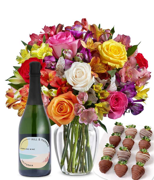 A bouquet of colorful roses and alstroemeria in a clear vase is displayed beside a bottle of Hill &amp; Harbor Sparkling Wine and 12 chocolate-covered strawberries.
