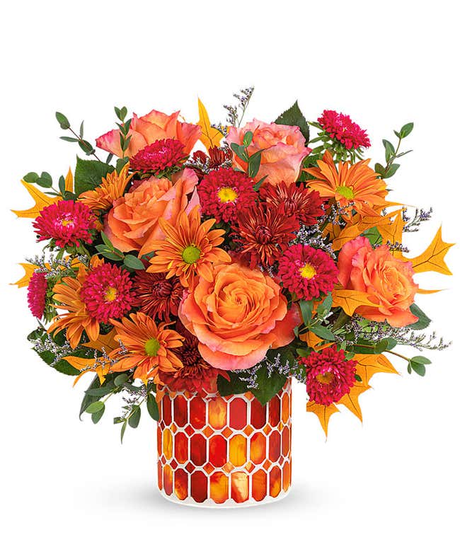 A vibrant floral arrangement featuring orange roses, red Matsumoto asters, bronze mums, and bronze daisies, complemented by greenery and fall leaves. The arrangement includes a personal card message and is elegantly presented in a keepsake vase&amp;#8203;